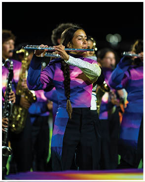 A person with a long braid wearing a colorful uniform is playing a flute in a marching band at night. Description generated by AI
