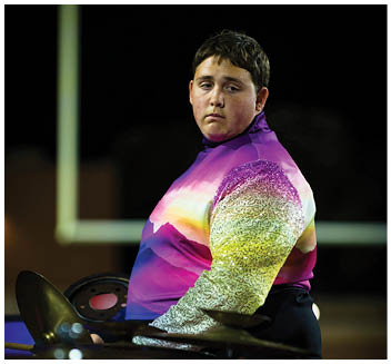 A person wearing a colorful, gradient long-sleeve shirt is standing behind a drum set, looking slightly to the side in a dimly lit environment. Description generated by AI