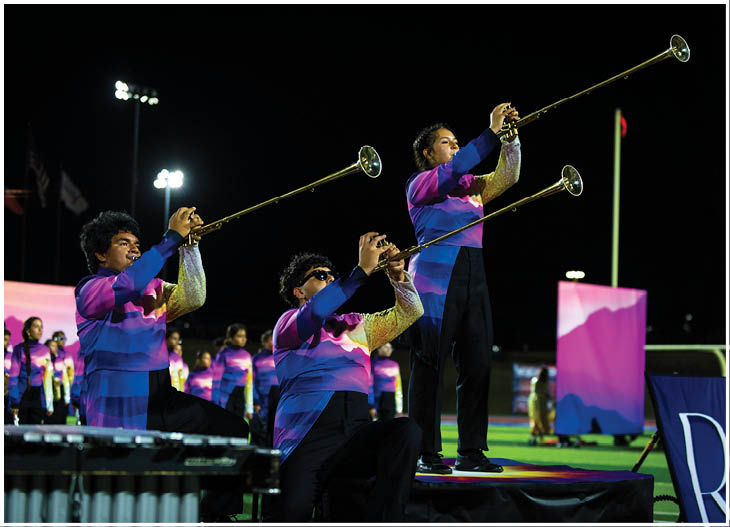 Three people wearing colorful uniforms are playing long brass trumpets on a stage at night with a group of similarly dressed people standing in the background. Description generated by AI