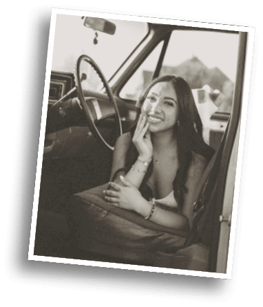 A person with long hair is smiling and resting their face on one hand while sitting inside a vintage car with a steering wheel visible. Description generated by AI