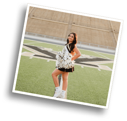 A person wearing a cheerleading uniform and holding pom-poms stands on a football field with empty bleachers in the background. Description generated by AI