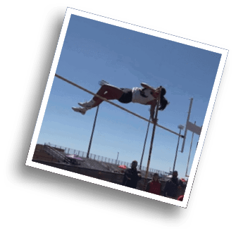 Two people stand near a pole vault setup on an outdoor track and field area with bleachers and a clear sky in the background. Description generated by AI
