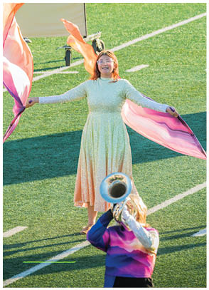 A person in a light-colored dress is performing with two flowing flags on a sports field while three people in colorful uniforms play brass instruments in front. Description generated by AI