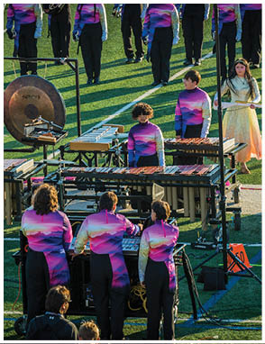 People wearing matching purple and pink gradient uniforms are playing percussion instruments on a field during a performance or rehearsal. Description generated by AI