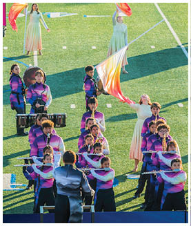 A group of people in colorful uniforms are performing a coordinated marching band routine on a grassy field with some holding flags and others playing instruments. Description generated by AI