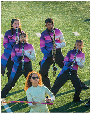People wearing colorful uniforms are playing clarinets on a grassy field while a person in a light-colored dress with curly hair is twirling a baton in front of them. Description generated by AI