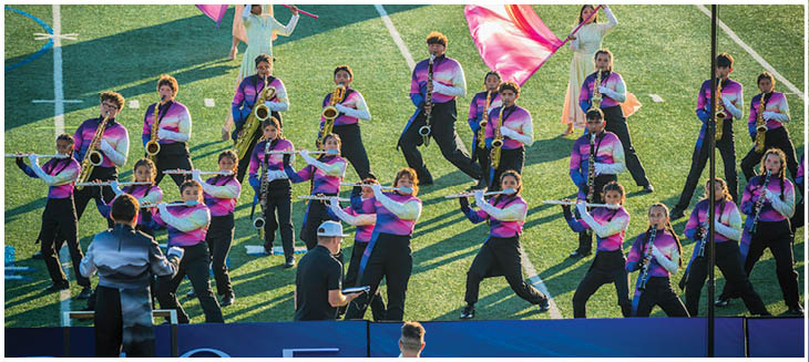 A group of people in colorful uniforms are playing musical instruments on a grassy field while two people stand in front of them, one holding a clipboard. Description generated by AI