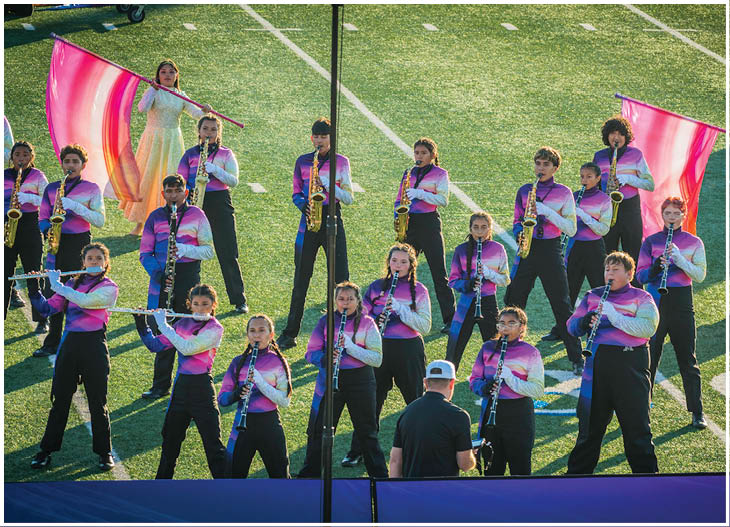 A marching band dressed in colorful gradient uniforms is performing on a football field with two people holding large pink flags. Description generated by AI