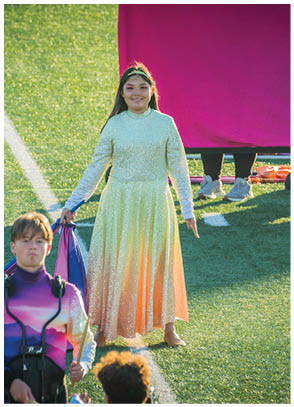 A person in a long, flowing dress stands on a field holding a flag while another person in a colorful uniform with a headset stands nearby. Description generated by AI