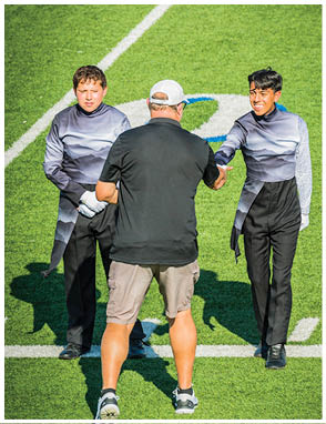 A person in a dark patterned outfit stands on a sports field next to another person wearing a black shirt and white cap facing away. Description generated by AI