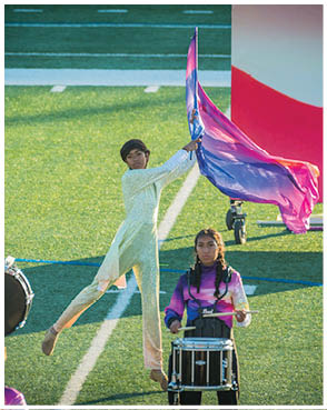 A person in a light-colored outfit is mid-air holding a flowing flag while another person in a tie-dye shirt plays a snare drum on a grassy field. Description generated by AI