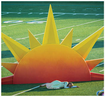 A person in a white outfit is lying on the ground in front of a large, colorful sun prop on a sports field with artificial turf. Description generated by AI