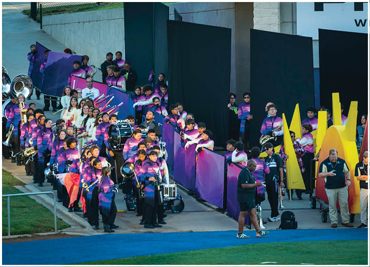 A large group of people in colorful uniforms are gathered outdoors with musical instruments and flags, appearing to prepare for a marching band performance. Description generated by AI