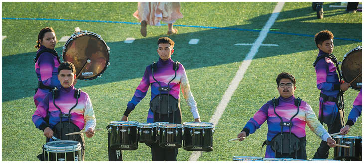 People wearing colorful uniforms are playing marching band drums and a saxophone on a grassy field. Description generated by AI