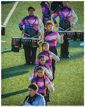 People in colorful uniforms are standing in formation on a grassy field, playing musical instruments including drums and a saxophone. Description generated by AI