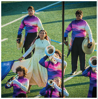 People in colorful uniforms are performing on a field with musical instruments and flags, with one person in a sparkling dress holding a flag. Description generated by AI
