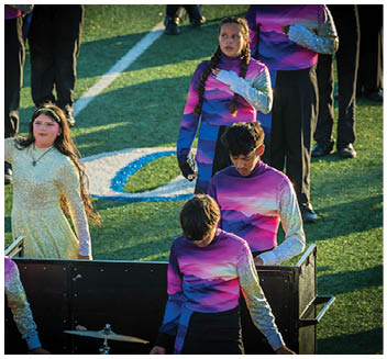 People wearing colorful performance uniforms stand on a field with one person in a sparkly dress and others in matching purple and silver outfits. Description generated by AI
