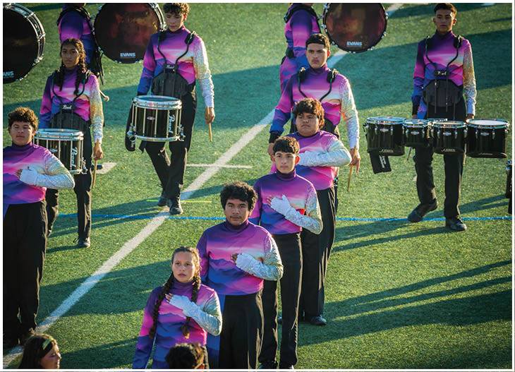 People wearing matching purple and pink gradient uniforms are standing on a field holding various marching band drums. Description generated by AI