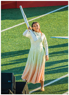 A person in a long light-colored dress stands on a field holding a flagpole while another person in a colorful top and dark pants plays a brass instrument behind them. Description generated by AI