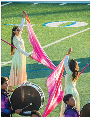 Adults in colorful uniforms are performing with drums and flags on a sports field during daylight. Description generated by AI