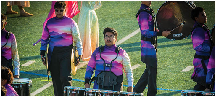 People wearing colorful uniforms are playing marching tenor drums on a grassy field. Description generated by AI