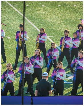 A group of people in matching purple and white uniforms are playing musical instruments on a field while two people in long dresses hold colorful flags behind them. Description generated by AI