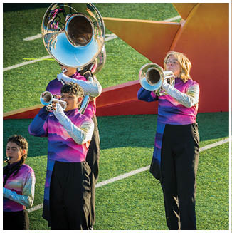 People wearing colorful uniforms are playing brass instruments on a grassy field near a large orange star prop while another person in a flowing yellow dress walks beside them. Description generated by AI