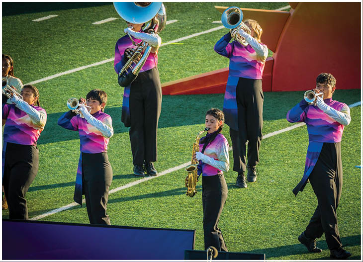 People in uniform pants are standing on a grassy field with a saxophone visible among them and a large screen or panel in the foreground. Description generated by AI