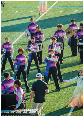 A marching band performs on a football field with people in colorful uniforms playing instruments and two people in flowing costumes dancing. Description generated by AI