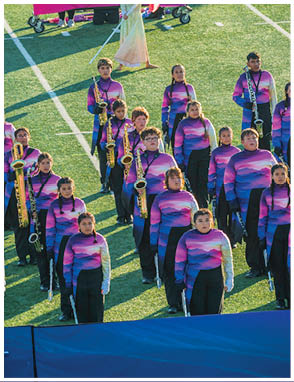 A group of people in matching purple and pink gradient uniforms stand on a football field with one person in a light-colored dress holding a large pink and purple gradient flag. Description generated by AI