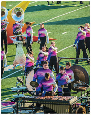 People in colorful uniforms perform with musical instruments and flags on a football field during a marching band show. Description generated by AI