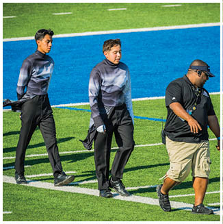 Three people walk on a green sports field with blue markings, two wearing matching black and gray uniforms and one wearing a black shirt, beige shorts, and a visor. Description generated by AI