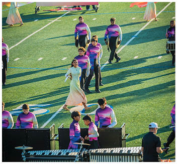 People in colorful uniforms and flowing dresses are performing a coordinated routine on a sports field with musical equipment visible in the foreground. Description generated by AI