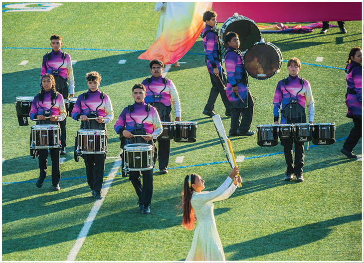 People in colorful uniforms are performing with drums and flags on a sports field during a marching band event. Description generated by AI