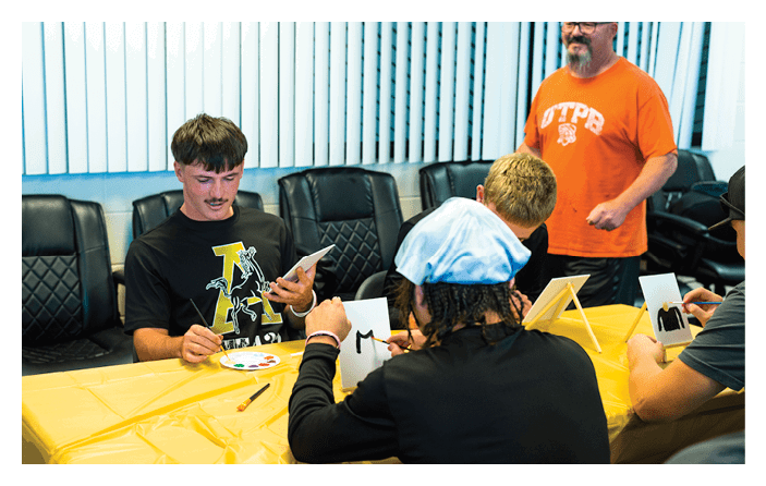 Several people are seated around a table covered with a yellow tablecloth, engaging in a painting activity with small canvases and paint palettes, while one person stands nearby wearing an orange shirt. Description generated by AI