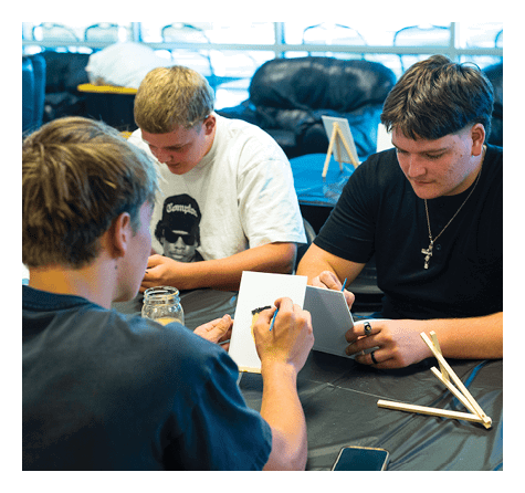 Three people with short hair sit around a table engaged in writing or drawing on small white cards in a casual indoor setting. Description generated by AI