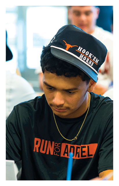 A person wearing a black shirt and a sideways cap with “Hook'em Horns" written on it is focused on writing or drawing on a small easel while sitting at a table with other people in a bright indoor setting. Description generated by AI