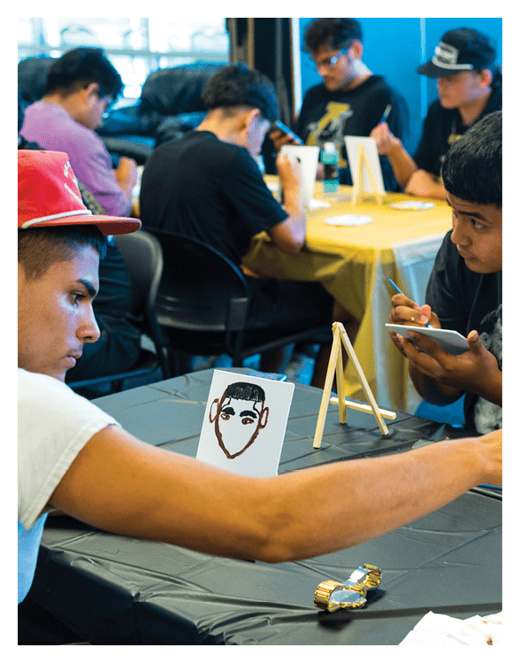 Two adults sit at a table engaged in painting, with one person holding a paintbrush and the other reaching for a jar of water, while a small drawing of a face is displayed on the table. Description generated by AI