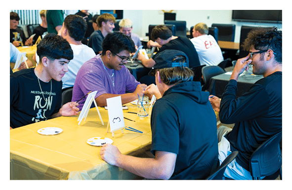 A group of people sitting around a table covered with a yellow tablecloth are engaged in a creative activity involving paint and small canvases. Description generated by AI