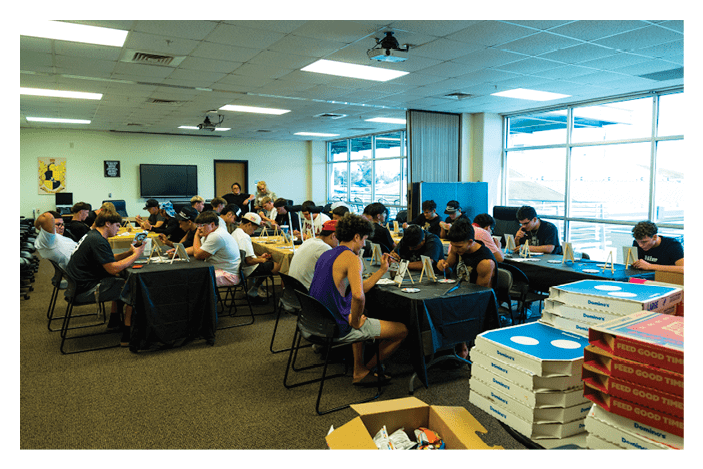 A group of people sitting at tables in a room engaged in activities with laptops and papers while several stacks of pizza boxes and an open box of snacks are visible in the foreground. Description generated by AI