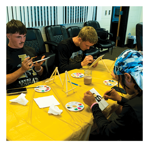 Three people sit around a table covered with a yellow cloth, focused on painting small canvases with brushes and paint palettes in a room with black chairs and vertical blinds. Description generated by AI