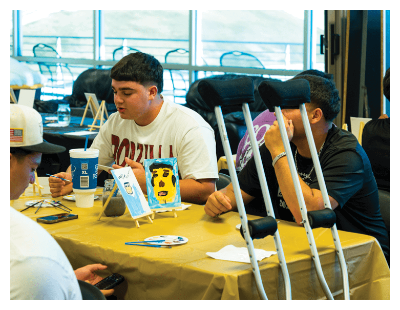 People sitting at a table covered with a yellow cloth are engaged in painting small canvases, with one person using crutches resting against the table. Description generated by AI