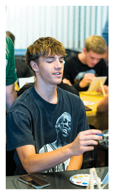 A person with short tousled hair wearing a black graphic t-shirt is sitting at a table holding a paintbrush with a palette of paint and a smartphone in front of them, while other people in the background are engaged in similar activities. Description generated by AI