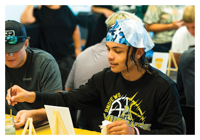 A person wearing a black shirt with a graphic design and a blue and white patterned headscarf is sitting and looking at something in front of them while other people are in the background. Description generated by AI