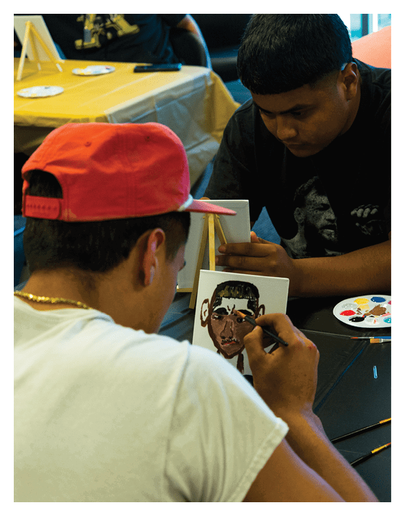 Two adults are seated at a table painting small portraits with brushes and paint palettes in a creative setting. Description generated by AI