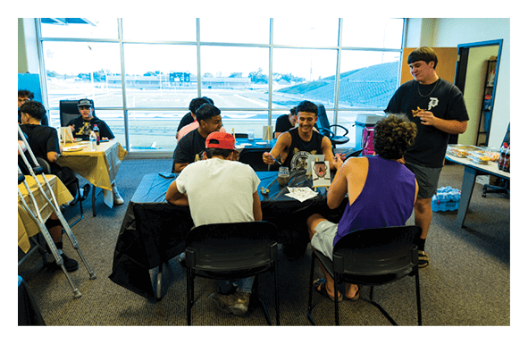 A group of people are gathered around tables in a room with large windows, engaging in conversation and activities, with some seated and one standing. Description generated by AI