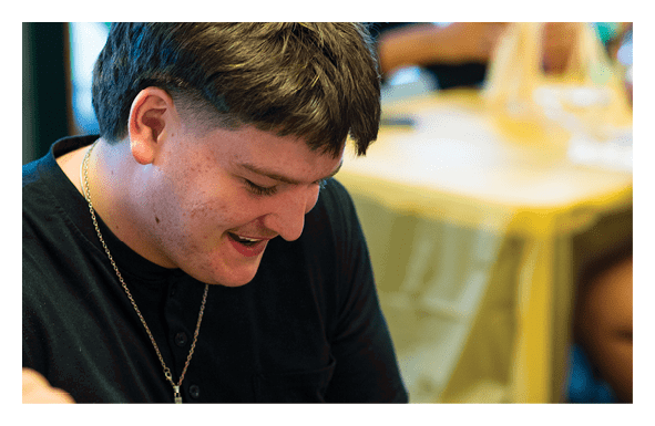 A person with short dark hair is looking down and smiling while two other people are blurred in the background sitting at a table. Description generated by AI