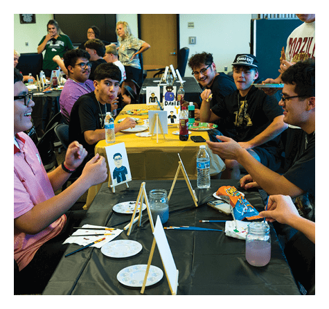 People sitting around a table engaged in a creative activity involving small easels, paintbrushes, and snacks. Description generated by AI