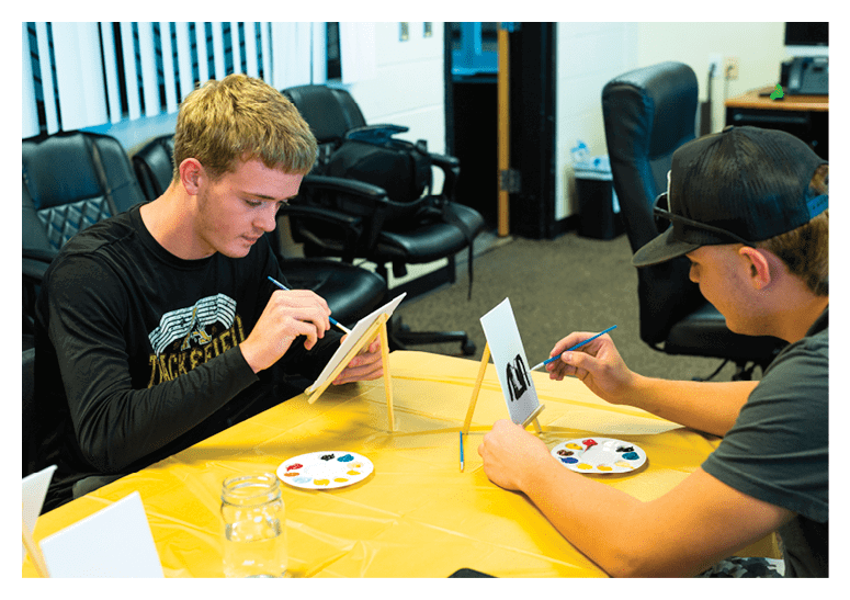 Two adults sit at a table covered with a yellow cloth, each painting on small canvases propped on easels, with paint palettes and brushes in front of them. Description generated by AI