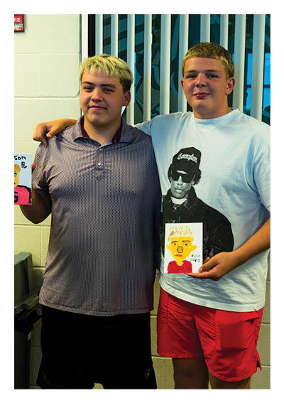 Two adults stand side by side indoors with one arm around the other while each holds a colorful drawing. Description generated by AI